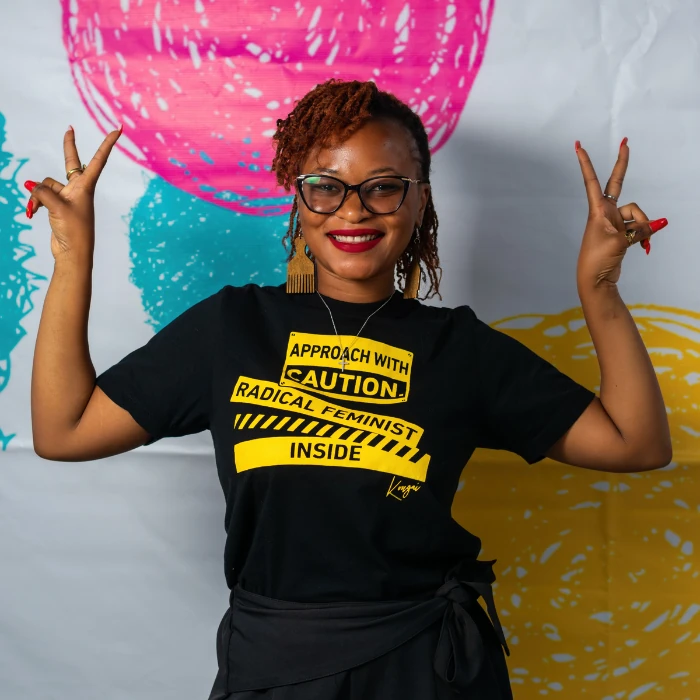 Chipasha Mwansa, vibrantly poses with both arms raised in two peace signs. Chipasha's black and yellow T-shirt reads: 'Approach with caution: Radical feminist inside'