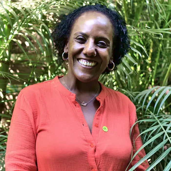 Wearing a red t-shirt, and with a multiplicity of green leaves in the background, Meron Estefanos looks radiantly into the camera.