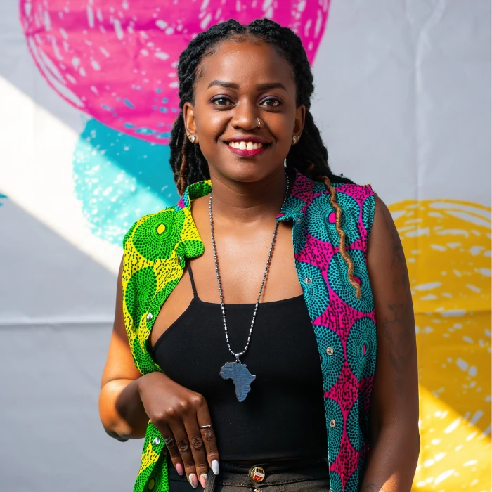 Sandra Kwikiriza wears a bright gilet in chitenge fabric, over a black strappy top and jeans, smiling widely into the camera.