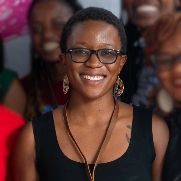Lena Anyuolo, sporting a short hair cut, wearing black-rimmed glasses and a bright smile looks directly into the camera. They wear a black tank top.